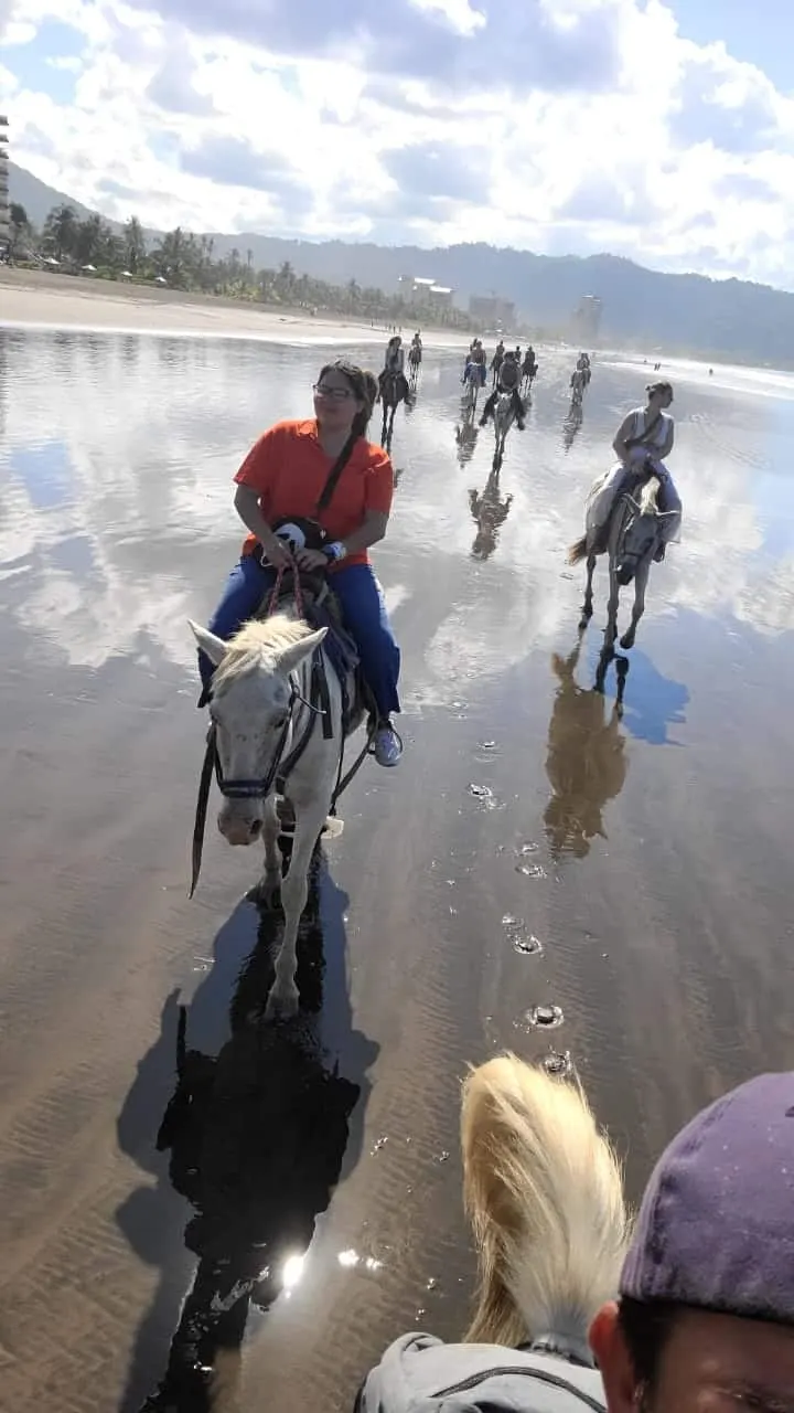 Horseback riders crossing the wide, reflective shoreline of Jacó Beach